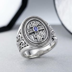 May include: A silver ring featuring an Orthodox cross design. The ring has an oval face with a detailed cross, a blue gemstone, and small accent stones. The band has an ornate, swirling pattern. The ring is set against a neutral background.