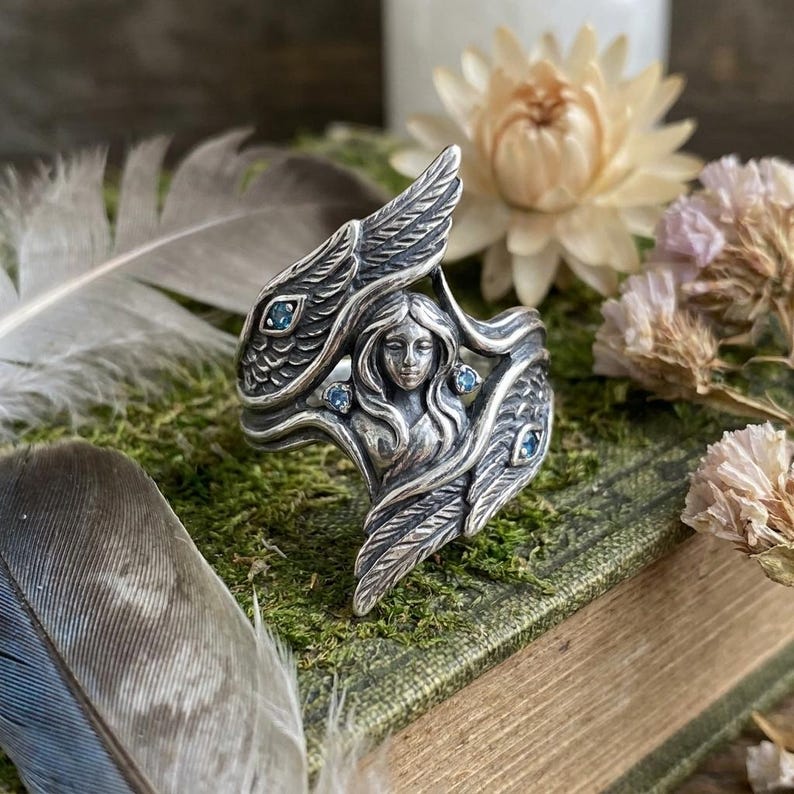May include: A silver ring featuring a central sculpted figure with flowing hair, flanked by two large textured wings. The design includes four small, round, light blue gemstones embedded within the wings and near the central figure's shoulders.