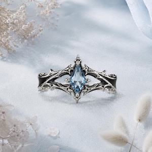 May include: A silver-toned ring featuring an intricate, pointed metal frame with black enamel detailing and a central, marquise-cut light blue gemstone. The design includes sharp, thorn-like accents extending from the center setting.