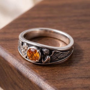 May include: A silver ring with an orange gemstone center. The ring features detailed angel figures with wings on the band. The ring is set on a wooden surface.