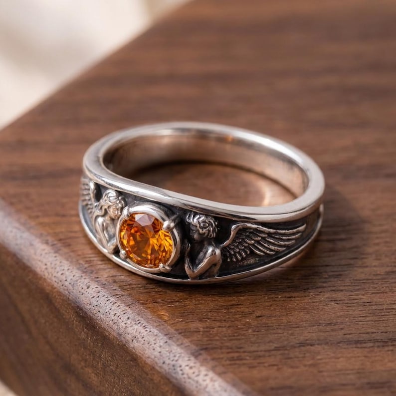 May include: A silver ring with an orange gemstone center. The ring features detailed angel figures with wings on the band. The ring is set on a wooden surface.