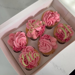 Puede incluir: Una caja de seis cupcakes con glaseado rosa y adornos dorados con purpurina. Los cupcakes están en una caja rosa claro con una ventana transparente. Los cupcakes tienen los nombres "Ella" y "Hello".
