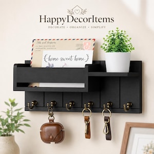 May include: Black wooden wall-mounted organizer with a shelf, mail slot, and six hooks. Includes a small potted plant, keys, and a brown leather case. The text "HappyDecorItems" and "home sweet home" are visible.
