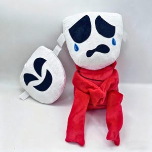Puede incluir: Un peluche con cabeza blanca, ojos negros tristes y detalles de lágrimas azules, unido a un cuerpo rojo con brazos largos. Un accesorio circular blanco separado con marcas negras curvas está colocado a la izquierda de la figura principal.