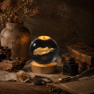 May include: A decorative glass sphere with a gold-colored cloud design, illuminated from within, sits on a wooden base. The sphere is surrounded by antique items, including a vase with dried flowers, an inkwell, and old books, creating a vintage aesthetic.