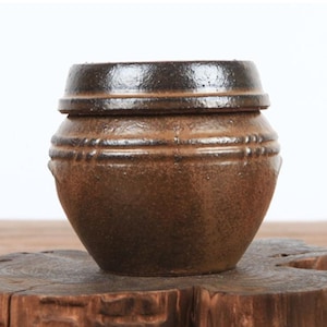May include: A small, round, brown ceramic jar with a dark, glossy rim and a matching lid. The body of the jar features a textured, mottled surface with two horizontal, raised bands encircling the upper portion, creating a rustic, earthy appearance.