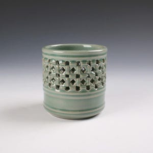 May include: A cylindrical ceramic vessel with a glossy sage green glaze. The upper section features a decorative band of cut-out diamond shapes and vertical bars, while the lower portion is smooth with two horizontal raised ridges near the base.