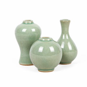 Puede incluir: Tres jarrones de cerámica color celadón de diferentes formas. Los jarrones tienen un esmalte craquelado y un acabado brillante. Las formas incluyen un jarrón bulboso, un jarrón redondo y un jarrón con cuello esbelto. Los jarrones se exhiben sobre un fondo blanco.