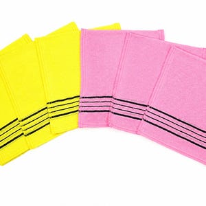 Puede incluir: Un conjunto de seis paños exfoliantes rectangulares dispuestos en forma de abanico. Tres paños son de color amarillo brillante y tres son de color rosa vibrante. Cada paño presenta una superficie texturizada y tres rayas negras horizontales.