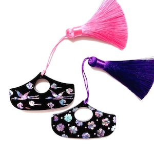 May include: Two black decorative pendants with a circular hole, adorned with mother-of-pearl accents. One features flying cranes and clouds, the other floral designs. Pink and purple tassels hang from the pendants, set against a white background.