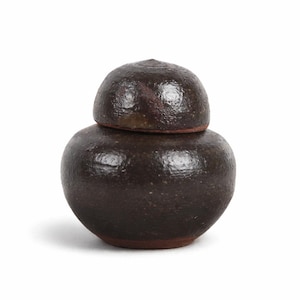 May include: A small, dark brown ceramic jar with a matching lid. The jar has a rounded body and a slightly textured surface. The lid sits snugly on top, creating a complete, decorative piece. The jar is likely handmade and has a rustic aesthetic.