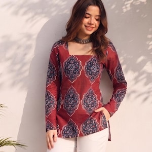 May include: A long-sleeved tunic featuring a deep red base with a repeating dark blue and white damask-style pattern, a square neckline, and side slits, paired with plain white trousers and a metallic choker necklace.