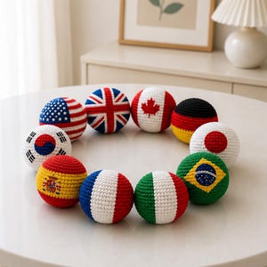 Puede incluir: Una colección de bolas de ganchillo, cada una con un diseño de bandera nacional diferente. Las banderas incluyen las de Estados Unidos, Reino Unido, Canadá, Alemania, Japón, Corea del Sur, España, Francia, Italia y Brasil. Las bolas están dispuestas sobre una superficie blanca.