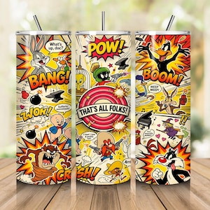 May include: A cylindrical tumbler featuring a colorful comic book collage of Looney Tunes characters including Bugs Bunny, Daffy Duck, and Tweety Bird. The design includes action words like POW, BANG, and BOOM, with the text That's All Folks in the center.