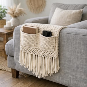 Puede incluir: Un organizador de reposabrazos de sofá tejido en color crema con dos bolsillos rectangulares, un patrón decorativo de macramé anudado y flecos largos. Los bolsillos contienen un libro de tapa dura marrón y un teléfono inteligente negro.