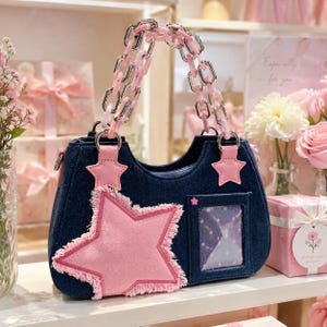 May include: A dark blue denim handbag with a pink star applique and a clear pocket with star details. It has a pink chain handle and pink star accents. The bag is a dark blue colour.