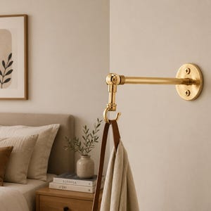 Op de afbeelding: Een aan de muur gemonteerde messing haak met een horizontale arm bevestigd aan een ronde basis met twee zichtbare schroeven. Een verticaal draaigewricht verbindt met een gebogen haak, die een bruine leren riem met een beige stoffen item draagt.