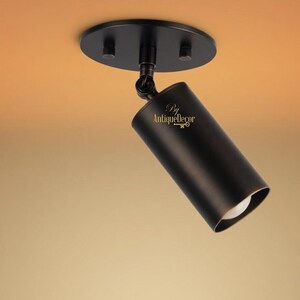 May include: A black ceiling spotlight with a cylindrical lamp head and a circular mounting plate. The light is angled downwards, revealing a white bulb. The fixture is mounted on a warm-toned ceiling.