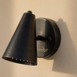 May include: A black wall sconce featuring a cone-shaped shade and a circular backplate. The shade has small perforations near the base. The text "by AntiqueDecor" is on the backplate. This light fixture is for interior use.