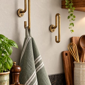 May include: Two wall-mounted brass hooks with a vertical, elongated U-shaped design, each secured by a circular base with three visible screws. A sage green kitchen towel with white horizontal stripes hangs from the left hook.