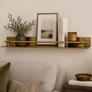 Può includere: Una mensola da parete in ottone con una barra orizzontale, che sostiene un vaso grigio scuro con fogliame verde, una fotografia di paesaggio in bianco e nero incorniciata, due libri bianchi intitolati Kinfolk e Live Beautiful e una candela in vetro marrone.