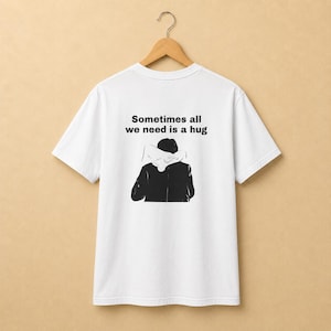 Puede incluir: Una camiseta blanca de manga corta con un estampado gráfico negro en la espalda. El gráfico muestra a una persona con una chaqueta oscura siendo abrazada por detrás, con el texto "Sometimes all we need is a hug" impreso en negro arriba.