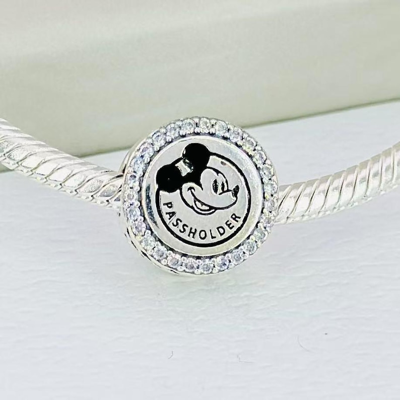 Pu&ograve; includere: Un ciondolo in argento con design circolare, incisione centrale di Mickey Mouse e la scritta PASSHOLDER. Il ciondolo &egrave; bordato da un anello di piccole pietre chiare e scintillanti ed &egrave; infilato su un bracciale a catena a serpente.