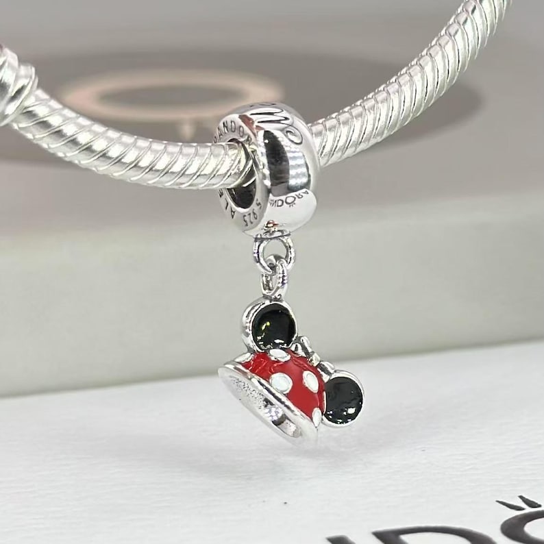 May include: A silver charm bead featuring a dangling Minnie Mouse hat design with red enamel, white polka dots, and black ears. The charm is attached to a silver snake chain bracelet. The metal surface is engraved with the text PANDORA and S 925.