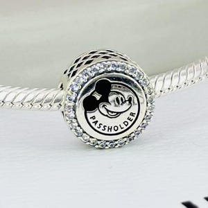 Pu&ograve; includere: Un charm in argento con un centro circolare che presenta una silhouette nera di Mickey Mouse e la scritta PASSHOLDER. Il charm &egrave; circondato da un bordo di piccole pietre trasparenti ed &egrave; infilato su un bracciale a catena snake in argento.