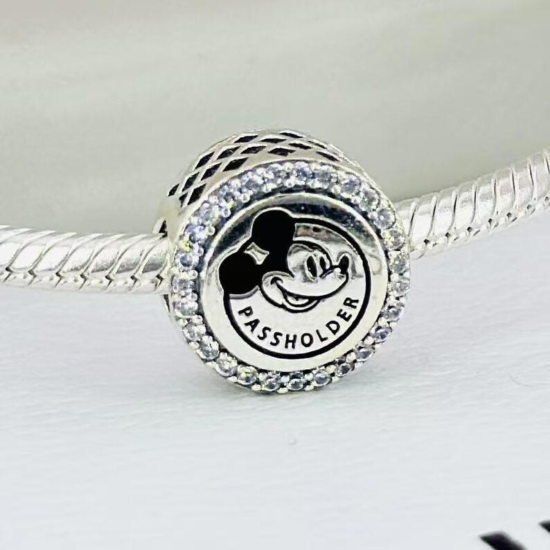 Pu&ograve; includere: Un charm in argento con un centro circolare che presenta una silhouette nera di Mickey Mouse e la scritta PASSHOLDER. Il charm &egrave; circondato da un bordo di piccole pietre trasparenti ed &egrave; infilato su un bracciale a catena snake in argento.