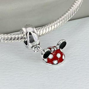 May include: A silver charm bracelet featuring a dangling pendant shaped like a red bow with black ears and white polka dots. The charm is attached to a polished silver snake chain with a smooth, rounded bail that connects to the main bracelet.