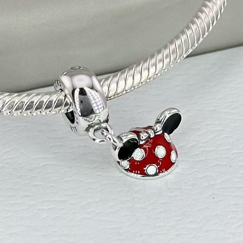 May include: A silver charm bracelet featuring a dangling pendant shaped like a red bow with black ears and white polka dots. The charm is attached to a polished silver snake chain with a smooth, rounded bail that connects to the main bracelet.