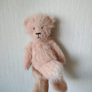 May include: A plush teddy bear with long, shaggy, light pink fur. The bear features dark, stitched eyes and a dark, stitched nose and mouth. The fur has a textured, slightly messy appearance, and the bear is shown in a seated position.