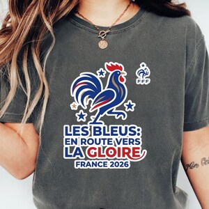 Pu&ograve; includere: T-shirt grigio scuro con grafica di gallo blu e rosso e la scritta "LES BLEUS: EN ROUTE VERS LA GLOIRE FRANCE 2026". La maglietta presenta anche il logo FFF e stelle.