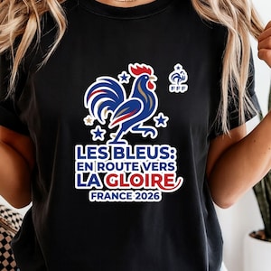 Pu&ograve; includere: T-shirt nero con un disegno di gallo blu, bianco e rosso e la scritta "LES BLEUS: EN ROUTE VERS LA GLOIRE FRANCE 2026". Include il logo FFF e stelle. Un capo a tema calcio francese.