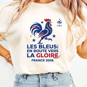 Pu&ograve; includere: T-shirt color crema con grafica di gallo blu, bianco e rosso, stelle e la scritta "LES BLEUS: EN ROUTE VERS LA GLOIRE FRANCE 2026". La maglietta include anche il logo FFF.
