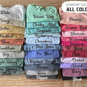 Pu&ograve; includere: Pile di magliette piegate in vari colori, tra cui bianco, avorio e pesca. Ogni maglietta &egrave; etichettata con il suo nome di colore, come "Island Reef" e "Black". Il testo "COMFORT COLORS ALL COLORS" &egrave; in alto.