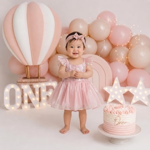 May include: A baby girl in a pink dress stands before a backdrop with balloons, a hot air balloon, and a cake. The word "ONE" is lit up in front of the baby. The cake has a "One" cake topper.