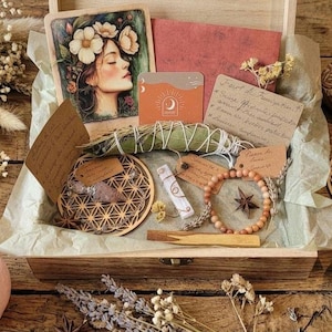 Op de afbeelding: Een houten kist gevuld met spirituele items, waaronder een bloemenkunstprint, saliebundel, houten armband en een rozenkwartskogel. Andere items zijn een kandelaar, houten onderzetters en gedroogde bloemen. De items zijn gerangschikt op een houten oppervlak.