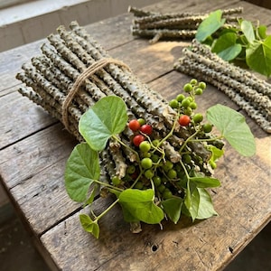 Könnte beinhalten: Ein Bündel aus holzigen, strukturierten Stängeln, die mit dünnem Bindfaden zusammengebunden sind, mit herzförmigen grünen Blättern und Trauben kleiner, runder grüner Beeren, durchsetzt mit einigen leuchtend roten Beeren, neben weiteren losen Stängeln.
