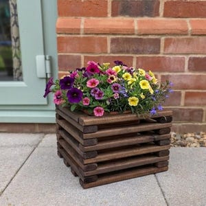 May include: A wooden planter box filled with vibrant flowers in shades of purple, pink, and yellow. The planter is constructed from horizontal wooden slats. It's placed on a grey stone patio, with a brick wall visible behind it.