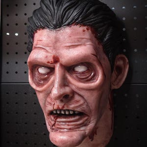 May include: A realistic, life-sized mask depicting a pale, weathered face with deep-set, solid white eyes, dark hair, and visible skin textures. The mask features blood-like red stains on the forehead, cheek, and chin, with a grimacing mouth.