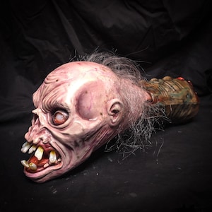 May include: A pale, textured creature head sculpture with a wide, open mouth revealing yellowed, uneven teeth. The figure has sparse, thin hair on the scalp and a detailed, wrinkled facial structure. A dark, segmented, organic-looking neck extends backward.
