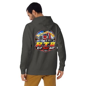 May include: A dark gray hooded sweatshirt featuring a circular graphic on the back. The design displays a red semi-truck on a road with a sunrise background, accompanied by the yellow and red text R.T.B and the words RIP TEAR BUST below it.