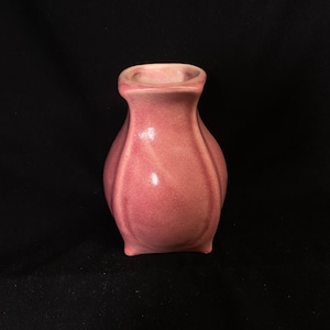 May include: A small, pink ceramic vase with a square base and a rounded body. The vase has a smooth, glossy finish.