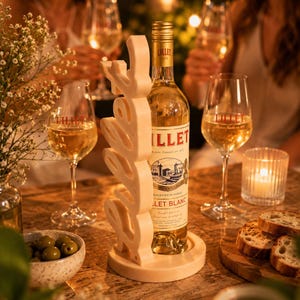 May include: A bottle of Lillet Blanc wine in a decorative beige wine holder, surrounded by wine glasses, olives, and bread. The wine holder has an abstract design. The scene is set on a wooden table, with soft lighting.