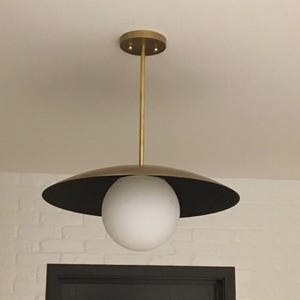 Peut inclure: Un luminaire suspendu composé d'un globe central en verre dépoli blanc suspendu à une tige métallique couleur laiton. Le globe est placé sous un large abat-jour circulaire plat couleur laiton avec un dessous noir mat foncé.