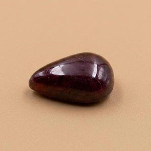 May include: A polished, teardrop-shaped gemstone in a deep burgundy color. The smooth surface reflects light, highlighting its rich hue. The stone is a natural mineral, likely used for jewelry or decorative purposes.