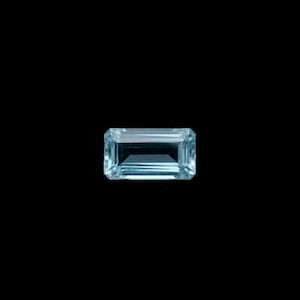 May include: A rectangular, emerald-cut gemstone with a translucent, pale light blue color. The stone features sharp, clean edges and a faceted surface that reflects light, creating a clear, crystalline appearance against a solid black background.