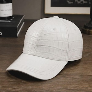 May include: A white baseball cap featuring a textured crocodile skin pattern across the crown and brim. The cap has a structured six-panel design with a top button, stitched seams, and eyelets for ventilation, presenting a clean and uniform look.
